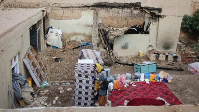 نمای یک خانه آسیب دیده. آوار به روی حویلی فرو ریخته. دو کودک در صحن حویلی ایستاده اند.