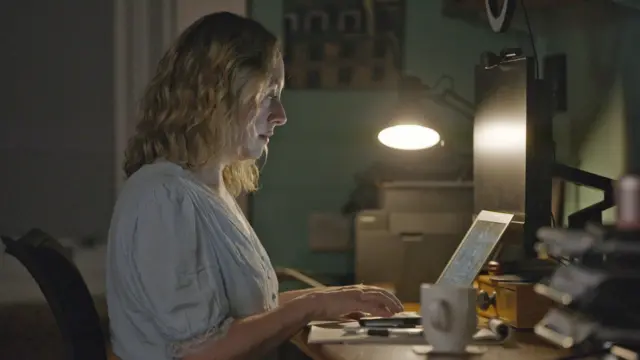 A TV grab of Catriona Stewart. She is siiting in a home office writing at a laptop with a desk light shining on the table. 