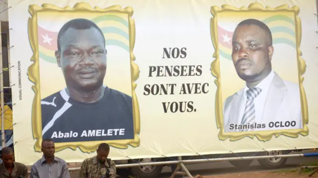 Une grande banderole affichant les portraits d'Abalo Amelete et de Stanislas Ocloo dans des cadres avec le message « Nos pensées sont avec vous » est exposée lors d'un enterrement, avec trois hommes assis devant dans le coin inférieur gauche de la photo.