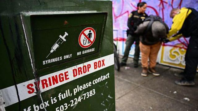 Dois policiais revistam um suspeito de tráfico de drogas em Portland, Oregon, ao lado de um lixo específico para o descarte de seringas