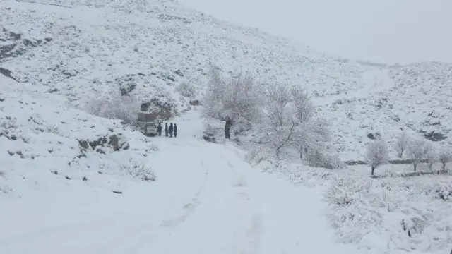 یک مسیر در طول کوتل که برف پوش است و یک موتر کوچک مسافربری و چند مسافر ایستاده در کنار موتر قرار دارند.