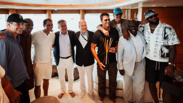 Un groupe de neuf hommes, dont Marcelo Claure, Didier Drogba, Rafael Nadal, le Prince Albert II de Monaco, Tom Brady, Marc Anthony et LeBron James, posent enlacés pour une photo. Ils se tiennent dans l'intérieur luxueux d'un bateau, orné de placages de bois et de tissus d'ameublement, tandis que Tom Brady tient un grand trophée composé de plusieurs pièces métalliques verticales.