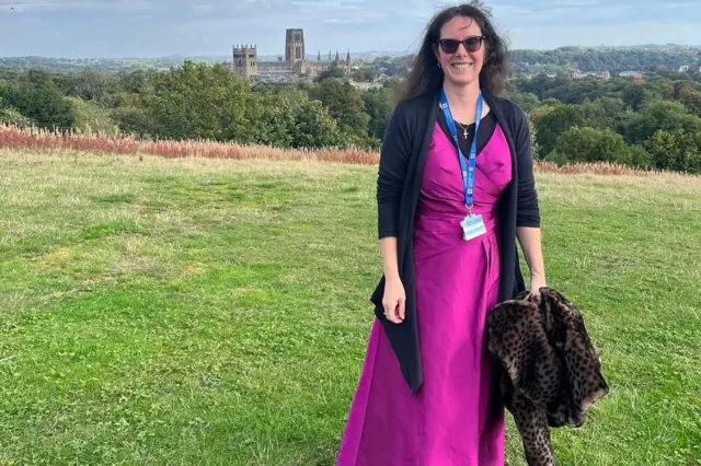 Jennifer Gane lleva un vestido de gala rosa oscuro hasta el suelo y un cárdigan negro. Está de pie en una colina con vistas a la Catedral de Durham.