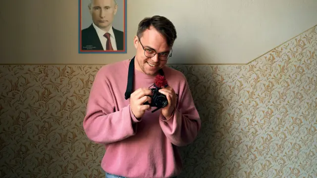 A man in a pink sweater holds a camera in front of a picture of Vladimir Putin