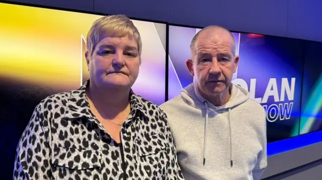 Linda tiene el pelo corto y rubio y lleva una camiseta con estampado de leopardo. George tiene el pelo muy corto y lleva una sudadera gris con capucha. Están de pie frente a televisores que dicen "The Nolan Show".