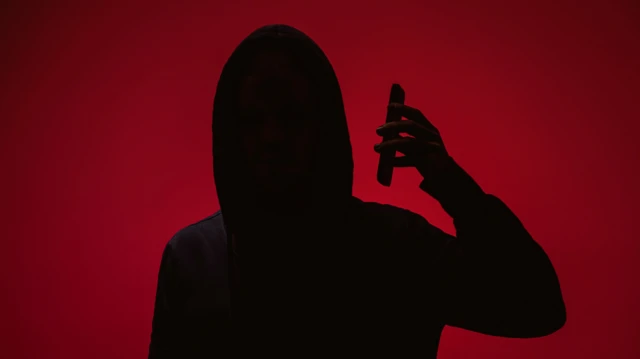 ظل شخص يرتدي قبعة رأس ويمسك هاتفاً محمولاً قرب أذنه، بخلفية حمراء موحّدة، مع إخفاء كامل لملامح الوجه