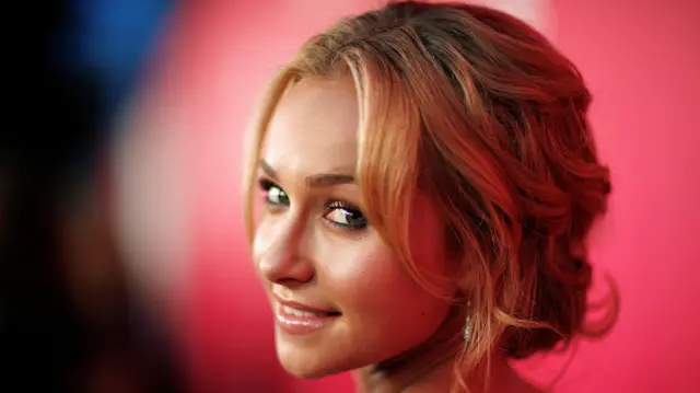 La cabeza de Hayden Panettiere se ve girada para mirar hacia atrás y sonreír suavemente, frente a un fondo rojo.