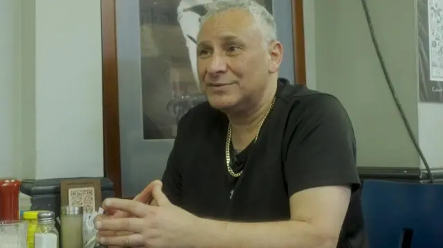 Se ve a Hussein vistiendo una camiseta negra y sentado en una mesa dentro de un café, con botellas de condimentos y menús en la mesa frente a él.