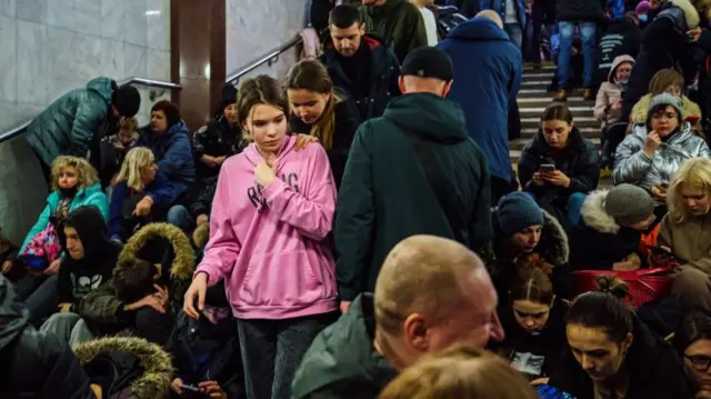 Une jeune fille vêtue d'un haut rose se tient dans une station de métro à Kharkiv, tandis que d'autres s'assoient autour d'elle.
