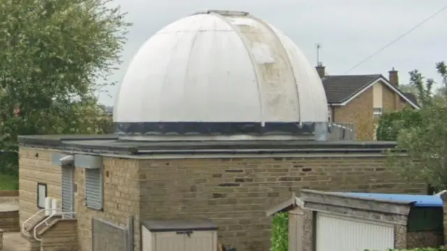 Un pequeño edificio de observatorio construido en ladrillo marrón claro con un techo plano y una gran cúpula blanca en la parte superior, que parece desgastada y ligeramente manchada.