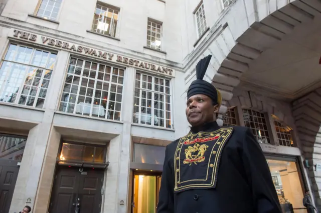 クリーム色の建物の外壁に、「ヴィーラスワミー・レストラン」の文字がある。手前には、胸元に「ヴィーラスワミー」と言う金色の文字が入った黒い服と、ターバンを巻いたようなデザインの黒い帽子を着けた男性が立っている