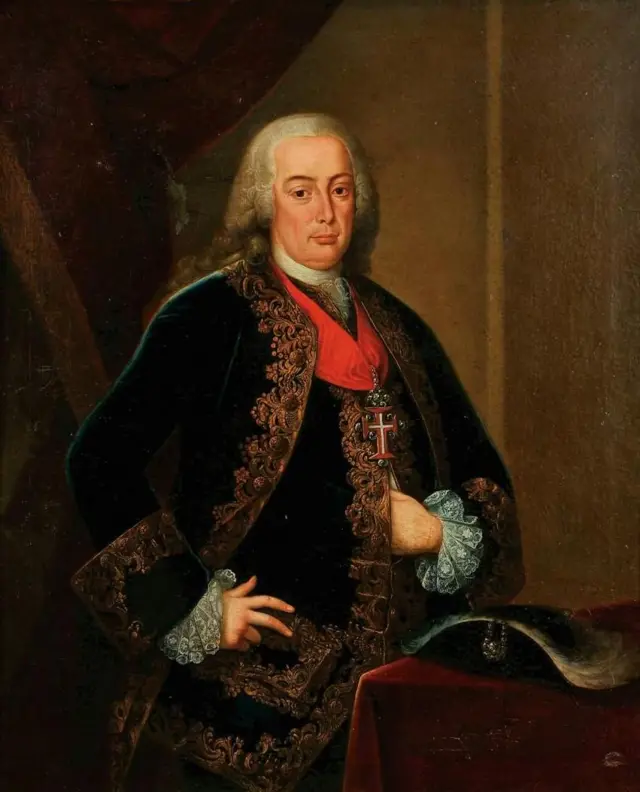 Retrato do Marquês de Pombal, pintado em 1766, mostrando-o de pé com expressão solene e postura confiante. Ele veste traje formal escuro com ricos bordados e um manto decorado. A mão direita repousa na cintura e a esquerda sobre uma mesa coberta por tecido vermelho, simbolizando autoridade e poder.
