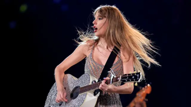 Taylor Swift em apresentação ao vivo, tocando um violão prateado coberto de brilho enquanto canta. Com os cabelos esvoaçando durante o movimento e vestindo um figurino prateado com franjas, ela aparece iluminada pelos refletores do palco.