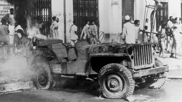 সাদা কালো ছবিতে একটি গাড়ি থেকে ধোয়া বের হতে দেখা যাচ্ছে, পেছনে কলাপসিবল গেট লাগানো একটি দেয়ালের পাশ দিয়ে হেঁওট যাচ্ছে কিছু মানুষ