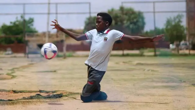 Zaher juega al fútbol con entusiasmo en un campo improvisado. El adolescente tiene los brazos abiertos mientras la pelota está en el aire a la altura de su pecho y él se apoya en el suelo con sus rodillas. Viste jeans gastados y una camiseta blanca.