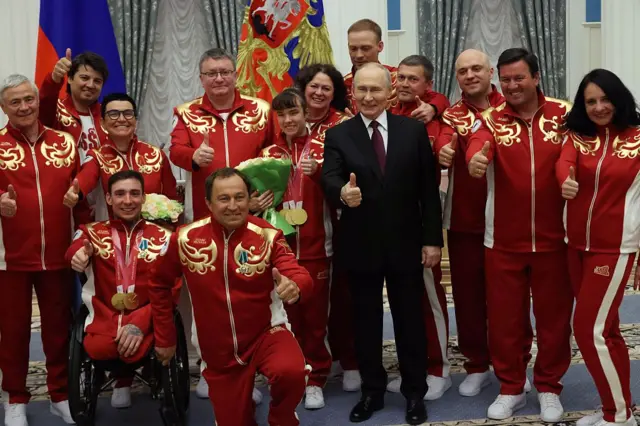 Група людей у червоних спортивних костюмах із золотистими візерунками, які позують для спільного знімка в урочистій залі. У центрі стоїть Путін в темному костюмі, а навколо – спортсмени та представники делегації, частина з яких демонструє піднятий великий палець. Дехто тримає медалі та букети квітів. На задньому плані видно прапор РФ та декоративний герб.