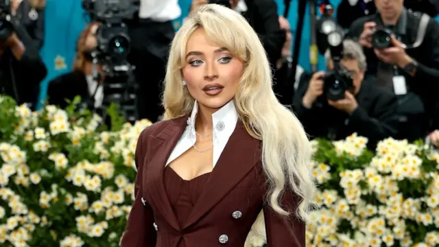 Sabrina Carpenter ma na sobie skrojoną bordową marynarkę z dużymi klapami i srebrnymi guzikami, a blond fale włosów opadają jej na ramiona — za nią widać grupę fotografów.