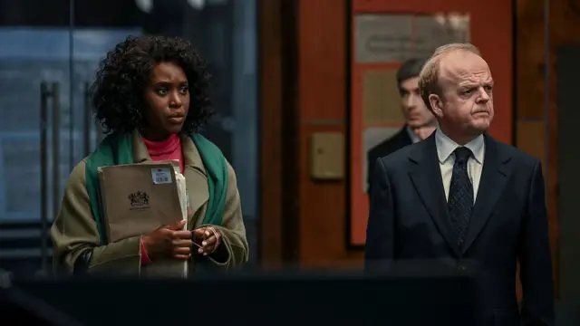 Кадр із фільму - чоловік та жінка стоять у приміщенні і дивляться вбік, жінка тримає в руках папку з документами