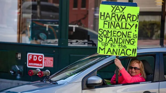 Женщина в машине с плакатом (на английском) «Ура, Гарвард! Наконец-то! Кто-то противостоит маньяку!»