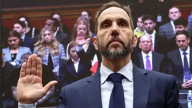 立ち上がり、右手をあげて宣誓するスーツ姿のスミス元特別検察官。その様子が、背後のモニターに映っている。スミス氏の後ろには他の証人や関係者が4列にわたり座っている