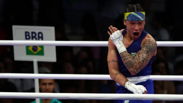 Bia é uma mulher negra, usando um shorts azul, camiseta regata azul, uma bandana com a bandeira do Brasil, no ringuecaça níquel a era do geloboxe comemorando.