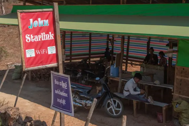 တာပေါ်လင်စတွေနဲ့ ဆောက်ထားတဲ့ စားသောက်ဆိုင်နဲ့ တွဲဖက်ဖွင့်ထားတဲ့ စတားလင့်ခ်အင် တာနက်ဆိုင်မှာ အင်တာနက်သုံးနေတဲ့ အမျိုးသားတစ်ဦးနဲ့ စားပွဲတစ်ခုနောက်မှာ မတ်တပ်ရပ်နေတဲ့ အမျိုးသမီး ၂ ဦး 