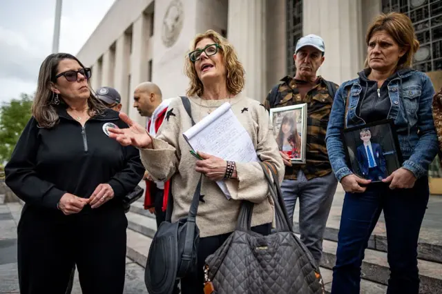 Fotoğrafta altı kişi var. Üç kadın ve bir erkek ön planda. Kadınlardan biri elinde not defteri ile konuşuyor. Sol tarafında siyahlı bir kadın onu dinliyor. Sağ tarafında bir kadın ve bir erkek ellerinde çocuk fotoğrafları taşıyor ve kadını dinliyor. 