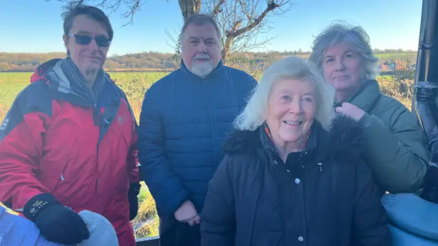 Una imagen de cuatro personas de pie afuera con un campo detrás. A la izquierda de la imagen hay dos hombres, a la derecha dos mujeres. El hombre en el extremo izquierdo lleva gafas de sol, un impermeable rojo y guantes gruesos. El hombre a su lado tiene barba y bigote blancos y viste un abrigo azul. La mujer en el extremo derecho viste un abrigo verde claro. Frente a ella hay una mujer con el pelo blanco ligeramente más largo y un abrigo azul oscuro.