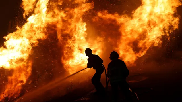 Двое пожарных тщетно пытаются залить бушующий лесной пожар