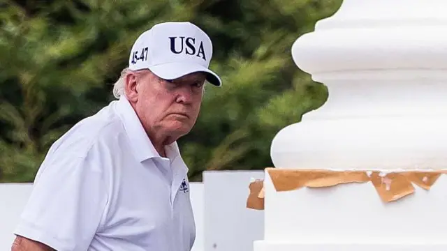 Donald Trump, vestido con polo blanco, pantalón negro y una gorra blanca con el lema "USA", junto a una gran columna blanca. 