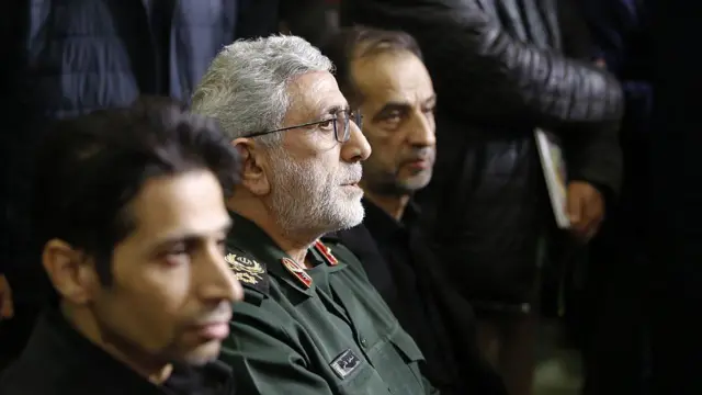 Tuğgeneral İsmail Kani, 17 Şubat 2026'da düzenlenen bir anma töreninde, iki adamın arasında, sağa dönük bir şekilde oturuyor ve yüzünü aydınlatan bir ışık huzmesi görülüyor.