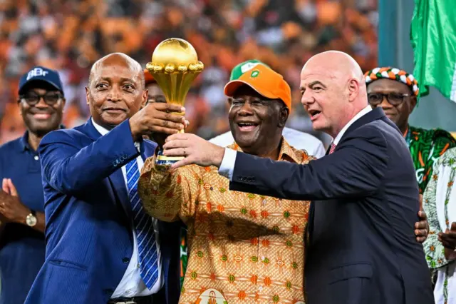 Le président de la Confédération africaine de football (CAF), Patrice Motsepe (à gauche), le président de la Côte d'Ivoire, Alassane Ouattara (au centre), et le président de la FIFA, Gianni Infantino (à droite), brandissent le trophée de la Coupe d'Afrique des Nations après la victoire de la Côte d'Ivoire en finale de la CAN 2024 contre le Nigeria, au stade olympique Alassane Ouattara d'Ebimpe, à Abidjan, le 11 février 2024.