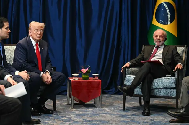 Trump e Lula sentados para coletiva de imprensa após encontro. Lula sorri