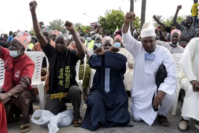 Trois hommes à genou les poings levés vers le ciel, l'un d'eux porte un -tshirt sur lequel est écrit : "Je suis Guinéen". Derrière eux une foule de gens.