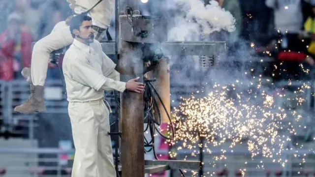 茶色い木の電柱のセットに、クリーム色の衣装を着たバッド・バニーがヘッドセットをつけて上っている。電柱からは火花が散り、白い煙が出ている。着ているフットボールジャージーに見えるトップスには白く「64」と描かれている