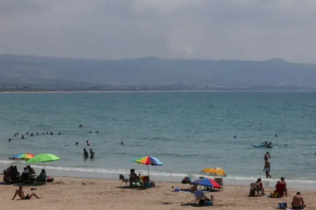 A imagem mostra uma praiaeu casinoum dia ensolarado com pessoas na areia e no mar e no fundo, depoiseu casinouma sérieeu casinomontanhas, uma fumaça. 