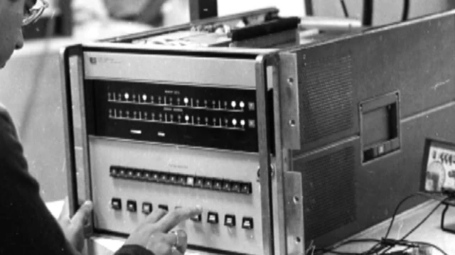 Fotografia em preto e branco mostra uma pessoa operando um computador antigo, com painel frontal repleto de indicadores luminosos, botões e controles, em um ambiente que parece ser um laboratório ou centro de pesquisa.