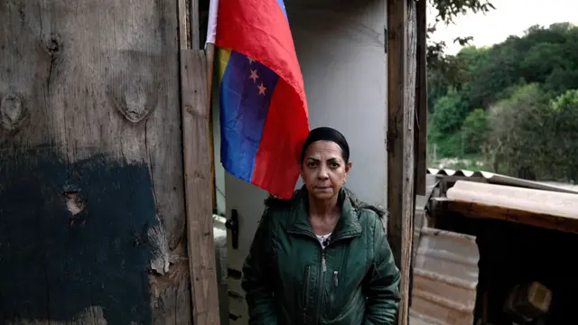 Yoraima Herrera, vestida com um casaco verde, em frente a seu barraco. Acima, uma bandeira da Venezuela pregada na madeira