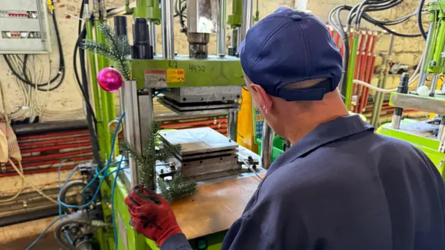 Homem de costas usa uma máquina para produzir decorações natalinas, com o ramo de uma árvore de Natal na mão e uma bola rosa pendurada na máquina