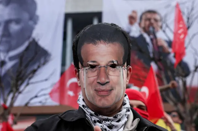 Seorang pengunjuk rasa mengenakan topeng Ekrem Imamoglu dengan lubang di matanya di depan bendera Turki dan spanduk dengan gambar Imamoglu