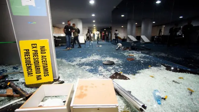Detalhe do Congresso Nacional com vidros quebrados no chão e um cartaz: "Não aceitamos ex presidiários na presidência!". 