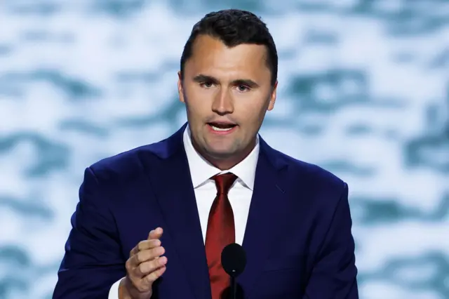 Charlie Kirk is standing behind a podium, wearing a dark blue suit with a white dress shirt and a shiny red tie. A black microphone is positioned in front of the podium. The background features a soft, out-of-focus pattern in light blue and white tones, creating a formal event or conference setting. One hand is raised slightly, suggesting he is speaking or gesturing during a presentation.