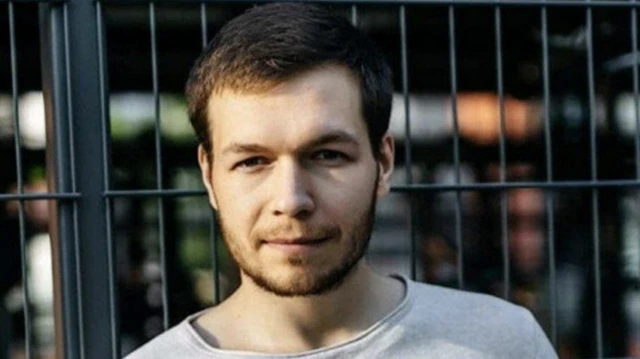 Александр Шишко, молодой человек с бородкой и усами, в серой футболке, стоит на фоне решетки и смотрит в камеру