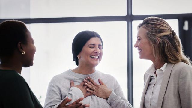 Duas mulheres brancas e uma negra conversam; ao centro, uma delas coloca as mãos sobre o peito em sinal de agradecimento e tem os olhos fechados; elas sorriem