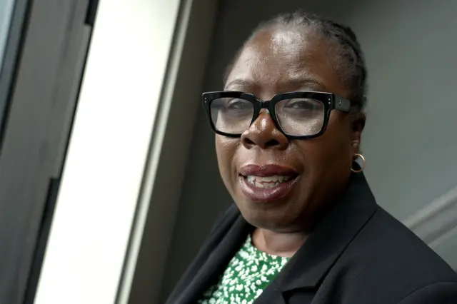 Портрет депутатки від Лейбористської партії Полетт Гамільтон. На ній чорні окуляри в товстій оправі, маленькі золоті кільця у вухах, а поверх зелено-білого топу з візерунком одягнений чорний блейзер.