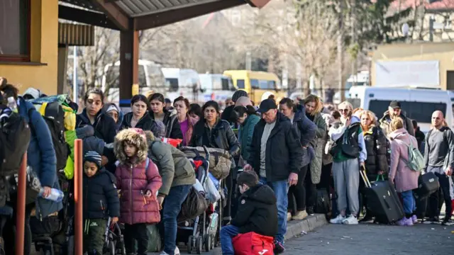 Mężczyźni, kobiety i dzieci w odzieży zimowej i z walizkami stoją w długiej kolejce. Na drugim planie widać płot i szereg zaparkowanych minibusów. 