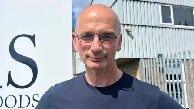 Un hombre con gafas sonríe a la cámara en una fotografía de hombros. Lleva una camiseta azul marino. Detrás de él se ve el exterior de una fábrica, con una valla metálica y el borde de un gran cartel blanco.