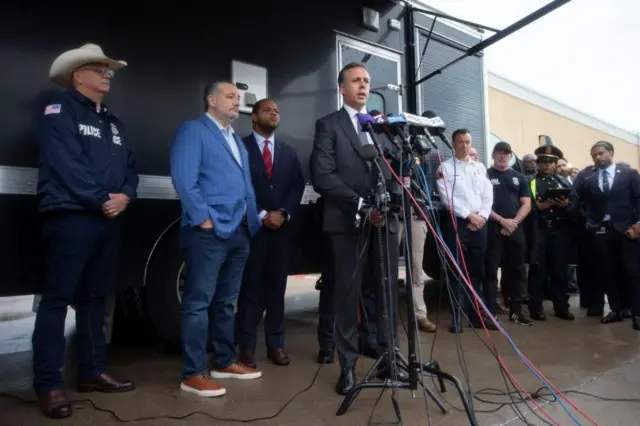 建物の外に置かれたマイクの前で、ダークスーツ姿の男性が話している。背後には、明るい青色のジャケットにジーンズ姿のクルーズ上院議員がいる。ほかにも関係者や警官が並んでいる