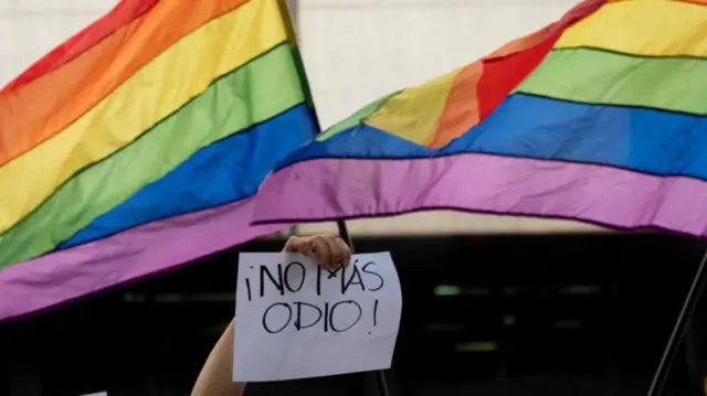 Mão com cartaz pedindo fim do ódio com bandeira do orgulho LGBT+ ao fundo