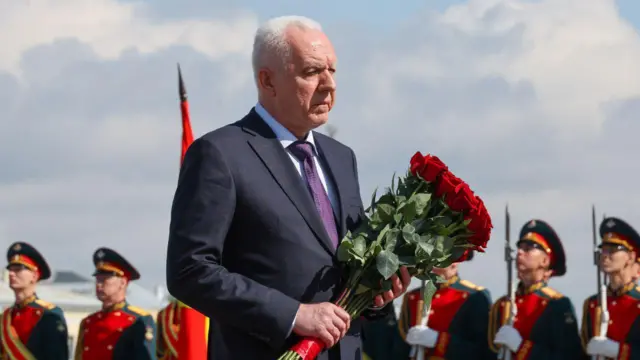 Пожилой седой мужчина в костюме с букетом цветов на фоне парадно одетых солдат со штыками на ружьях